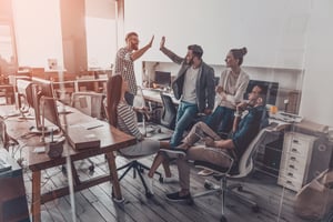 Two_cheerful_young_business_people_giving_high-five_while_their_colleagues_looking_at_them_and_smiling_